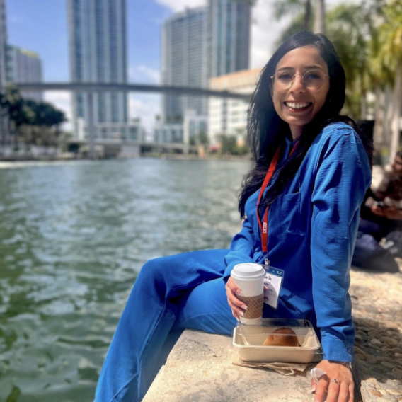 Giselle Caretto sitting on a stone wall by a river in a city, smiling.